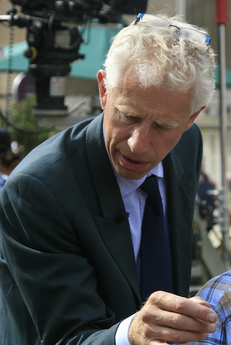 Rupert Maas speaking outdoors in a suit, leaning forward during an event.